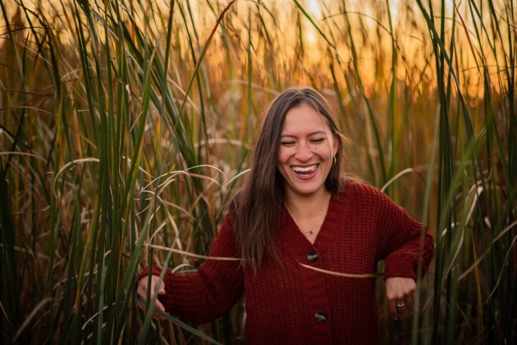 Woman with long brown hair and red sweater exiting tall reeds at sunset